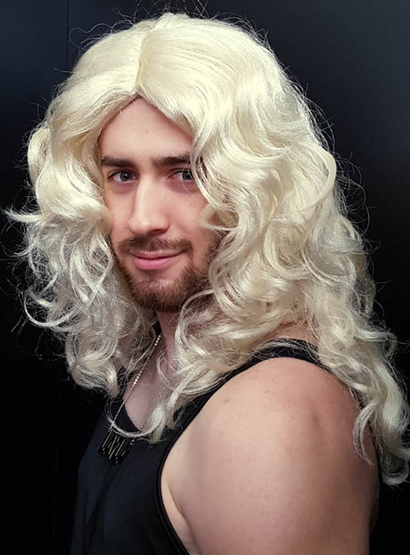 Eine Person mit hellbraunem Bart trägt die „Layla Perücke blond mit Locken“, eine lange, lockige, platinblonde Perücke, sowie ein schwarzes, ärmelloses Oberteil und steht vor einem schlichten, dunklen Hintergrund.