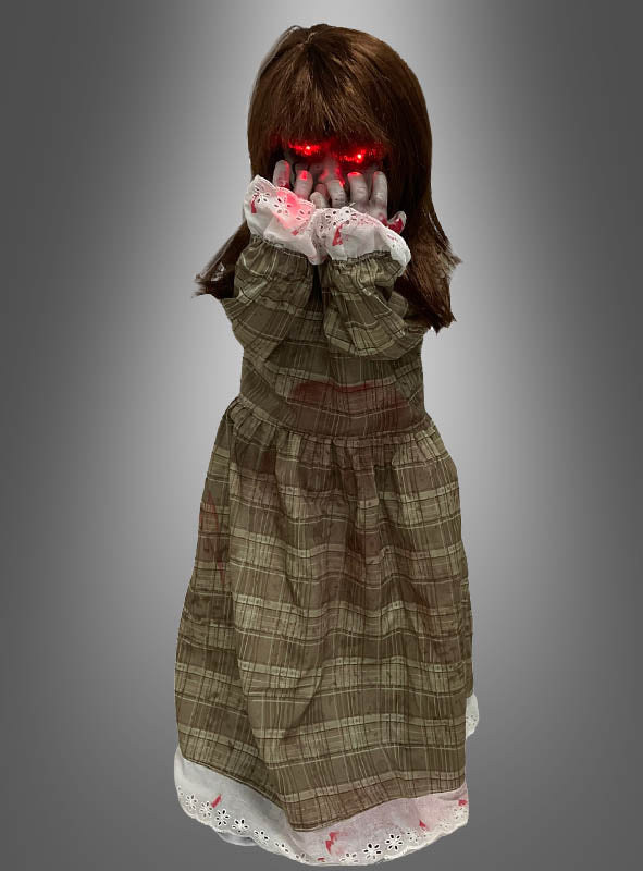 „Die Weinende Puppe, animiert, 90 cm“ ist eine Puppe mit braunen Haaren in einem karierten Kleid, die vor einem grauen Hintergrund ihr Gesicht mit leuchtend roten Händen bedeckt.