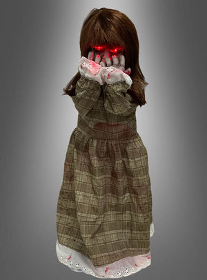 „Die Weinende Puppe, animiert, 90 cm“ ist eine Puppe mit braunen Haaren in einem karierten Kleid, die vor einem grauen Hintergrund ihr Gesicht mit leuchtend roten Händen bedeckt.