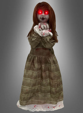 Die „Weinende Puppe“ (90 cm) hat leuchtend rote Augen, ein blutverschmiertes Gesicht und blutverschmierte Hände und trägt ein kariertes Kleid, während sie vor einem grauen Hintergrund steht.