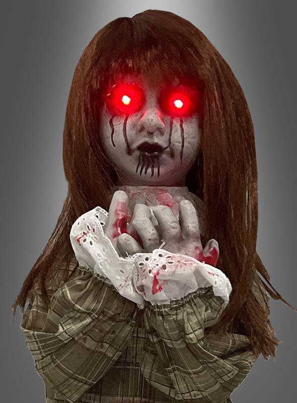 Die „Weinende Puppe“ (90 cm groß) hat langes braunes Haar, leuchtend rote Augen und schwarze Tränen. Sie trägt ein kariertes Outfit mit sichtbaren Blutflecken und hält die Hände vor dem Gesicht zusammen.