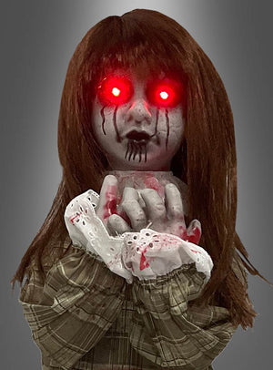 Die „Weinende Puppe“ (90 cm groß) hat langes braunes Haar, leuchtend rote Augen und schwarze Tränen. Sie trägt ein kariertes Outfit mit sichtbaren Blutflecken und hält die Hände vor dem Gesicht zusammen.