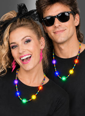 Zwei Personen in schwarzen T-Shirts, mit bunten Perlenketten wie der „Bunte LED-Partykette mit Licht“ und auffälligen Accessoires – darunter pinkfarbene Blitz-Ohrringe und dunkle Sonnenbrillen – lächeln und posieren vor einem schlichten Hintergrund.