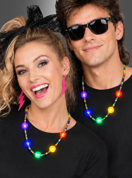 Zwei Personen in schwarzen T-Shirts, mit bunten Perlenketten wie der „Bunte LED-Partykette mit Licht“ und auffälligen Accessoires – darunter pinkfarbene Blitz-Ohrringe und dunkle Sonnenbrillen – lächeln und posieren vor einem schlichten Hintergrund.