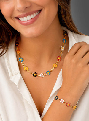 Eine Frau lächelt in einer weißen Bluse und trägt das „Buntes Blumenschmuck Set Armband & Kette“ – eine goldene Halskette und ein Armband, verziert mit bunten Emaille-Gänseblümchen –, das sich perfekt eignet, um jedem Outfit einen Hauch von Boho-Schmuck zu verleihen.