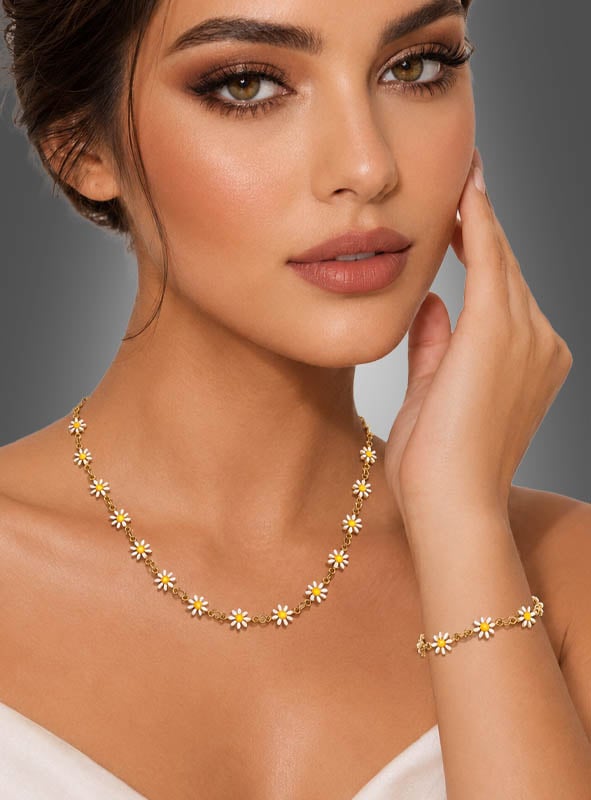 Eine Frau trägt das Schmuckset „Weiße Blumen“ – bestehend aus einer goldenen Gänseblümchen-Halskette und einem passenden Armband – kombiniert mit einem weißen, schulterfreien Oberteil, dezentem Make-up und sanft gewellten braunen Haaren. Vor einem grauen Hintergrund posiert sie und verkörpert damit einen mühelos schicken Boho-Schmuck-Look.