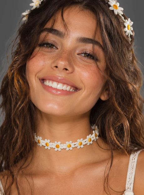 Eine lächelnde Frau mit welligem braunem Haar trägt ein Stirnband mit Gänseblümchen und das „Gänseblümchen-Halsband für Blumenkinder“ und blickt vor einem schlichten Hintergrund in die Kamera – ein perfektes Beispiel für den Boho-Accessoire- und Hippie-Schmuck-Stil.