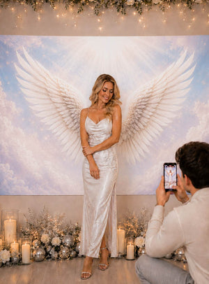 Eine Frau in einem weißen Kleid steht vor einer XXL-Fotowand mit der Wanddekoration „Weiße Engelsflügel“ (170 x 200 cm), während ein Mann kniet, um sie mit seinem Handy zu fotografieren. Umgeben von Kerzen und Blumendekorationen bildet die Szene einen idealen Hintergrund für ein Fotoshooting.