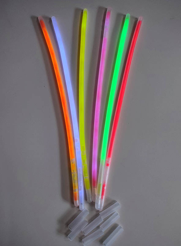 Sechs bunte Leuchtarmbänder aus der 100er-Packung – in Orange, Blau, Gelb, Grün und Pink – liegen auf einer grauen Oberfläche, daneben mehrere transparente Kunststoffverbinder. Die Leuchtarmbänder in der 100er-Packung sind das ideale Partyzubehör für jede Feier.