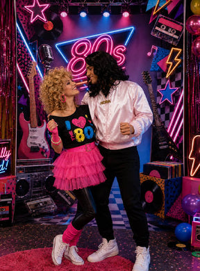 Zwei Personen in Kostümen im Stil der 1980er Jahre posieren gemeinsam in einem mit Neonlicht dekorierten Raum mit Gitarren, Schallplatten und farbenfrohem Retro-Dekor und zeigen ihre lustigsten Posen vor einem leuchtenden „80er-Party-Fotohintergrund XXL“ (160 x 220 cm) und einem Neon-Selfie-Stab.