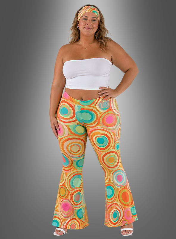 Model präsentiert die „Hippie Schlaghose Damen mit Stirnband“: Sie trägt die auffällige Schlaghose mit lebhaften kreisförmigen Mustern, ein passendes Stirnband, ein weißes Bandeau-Top und weiße Sandalen – und verkörpert so einen authentischen Retro-Look der 70er Jahre vor einem grauen Hintergrund mit Farbverlauf.
