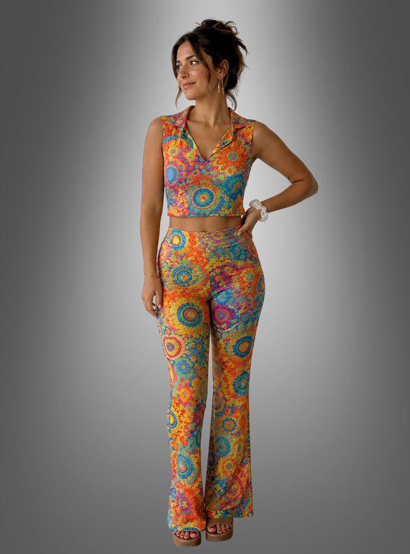 Eine Frau, die das „Buntes 70er Hippie Set Damen 2tlg.“ trägt – ein farbenfrohes, ärmelloses Crop-Top und eine dazu passende, hoch taillierte Schlaghose mit einem leuchtenden Mandala-Muster –, gestylt im klassischen Hippie-Look der 70er Jahre, steht vor einem grauen Hintergrund mit Farbverlauf.