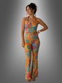 Eine Frau, die das „Buntes 70er Hippie Set Damen 2tlg.“ trägt – ein farbenfrohes, ärmelloses Crop-Top und eine dazu passende, hoch taillierte Schlaghose mit einem leuchtenden Mandala-Muster –, gestylt im klassischen Hippie-Look der 70er Jahre, steht vor einem grauen Hintergrund mit Farbverlauf.