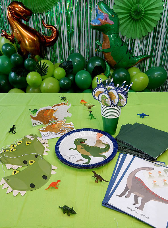 Der Tisch ist mit dem „Dino Party Deko Kindergeburtstag Set 81-teilig“ geschmückt, das Teller, Hüte, Luftballons, Servietten, Becher und kleine Dinosaurierfiguren zum Thema Dinosaurier enthält und durch grüne Luftschlangen und Luftballons im Hintergrund ergänzt wird.