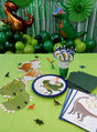 Der Tisch ist mit dem „Dino Party Deko Kindergeburtstag Set 81-teilig“ geschmückt, das Teller, Hüte, Luftballons, Servietten, Becher und kleine Dinosaurierfiguren zum Thema Dinosaurier enthält und durch grüne Luftschlangen und Luftballons im Hintergrund ergänzt wird.