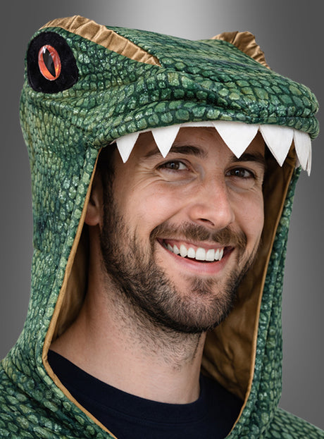 Ein bärtiger Mann lächelt, während er den grünen „Dino T-Rex Poncho für Erwachsene“ trägt, der wie ein Dinosaurier gestaltet ist und weiße Zähne sowie ein auffälliges rotes Auge an der Kapuze aufweist.