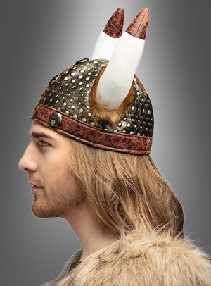 Eine Person im Profil, die einen Wikinger-Stoffhelm als Teil eines Nordmann-Kostüms trägt, ist in ein mit Pelz besetztes Outfit gekleidet und hat langes hellbraunes Haar; im Hintergrund ist ein grauer Farbverlauf zu sehen.