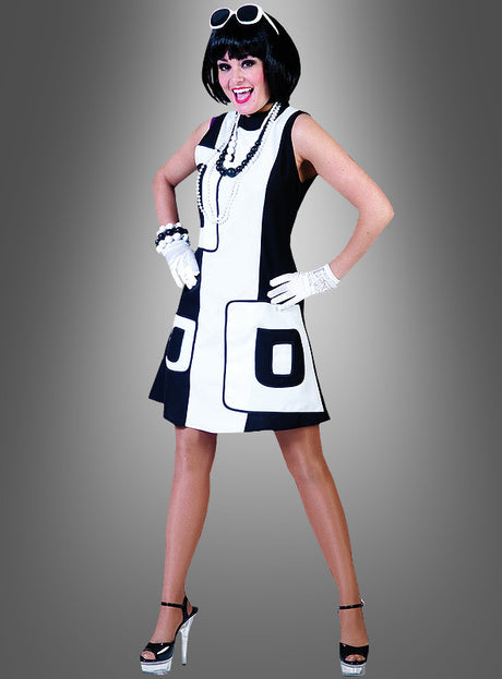 Eine Frau präsentiert das schwarz-weiße 60er-Jahre-Mod-Kleid, kombiniert mit weißen Handschuhen, auffälligem Schmuck und schwarzen Pumps, und posiert mit den Händen in den Hüften vor einem grauen Hintergrund mit Farbverlauf.