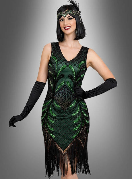Eine Frau, die das glamouröse Flapper-Kleid „Vivian“ in Grün und Schwarz mit Fransen trägt, kombiniert mit schwarzen Handschuhen und einem mit Perlen verzierten Stirnband, posiert vor einem grauen Hintergrund mit Farbverlauf.