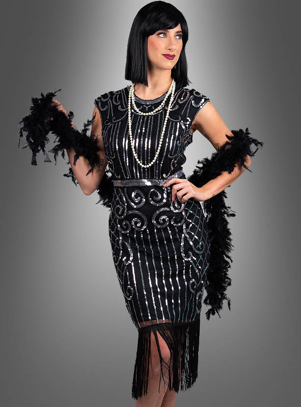 Eine Frau, die das „Premium 20er Jahre Kleider Kelly“ trägt – ein schwarzes Charleston-Kleid im Stil der 1920er Jahre mit Pailletten und Fransen –, kombiniert mit mehrreihigen Perlenketten, einer klassischen schwarzen Bob-Perücke und einer schwarzen Federboa, steht vor einem grauen Hintergrund: der Inbegriff glamouröser 20er-Jahre-Kleider.