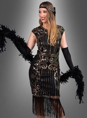Vor einem grauen Hintergrund steht eine Frau, die das „Premium 20er Jahre Kleider Kelly“ trägt – ein schwarz-goldenes Flapper-Kleid im Stil der 1920er Jahre, verziert mit Fransen, die an glamouröse Vintage-Mode erinnern –, dazu lange schwarze Handschuhe, ein Feder-Stirnband und eine schwarze Federboa in der Hand.