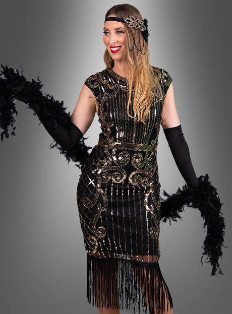 Vor einem grauen Hintergrund steht eine Frau, die das „Premium 20er Jahre Kleider Kelly“ trägt – ein schwarz-goldenes Flapper-Kleid im Stil der 1920er Jahre, verziert mit Fransen, die an glamouröse Vintage-Mode erinnern –, dazu lange schwarze Handschuhe, ein Feder-Stirnband und eine schwarze Federboa in der Hand.