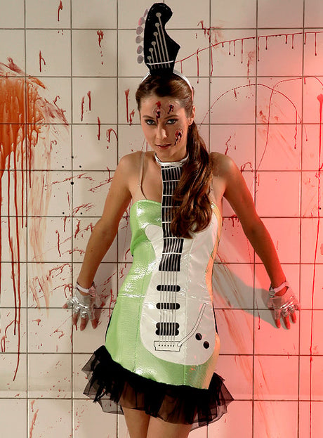 Eine Frau steht vor einer blutbespritzten Fliesenwand und trägt das „Disco 2. Wahl Gitarrenkostüm“ – ein glänzendes Kleid und einen Kopfschmuck mit Gitarrenmotiv –, dazu Halloween-Make-up und Handschuhe mit Skelettmuster.