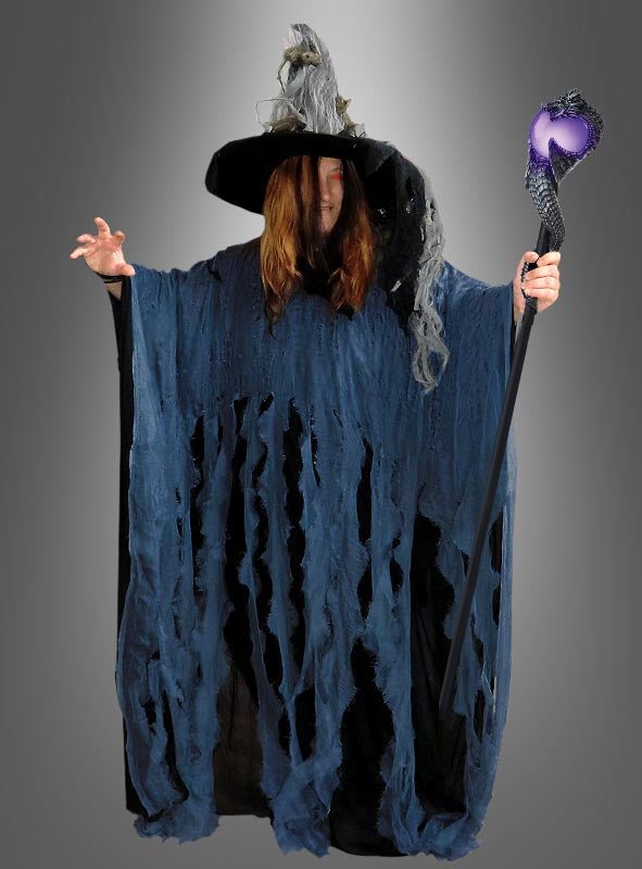 Eine Person, die einen zerfledderten Fetziger Überwurf-Poncho XXXL in Übergröße mit Kapuze und großem Hut trägt und einen Stab mit einem violetten Kristall an der Spitze in der Hand hält, steht vor einem grauen Hintergrund – ein auffälliges Hexenkostüm für Damen und Herren.