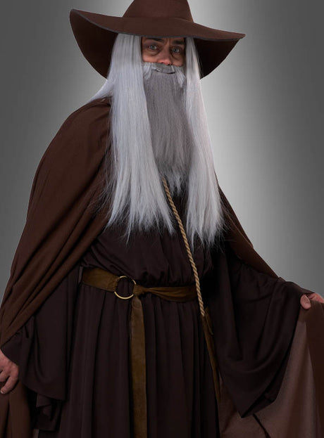 Eine Person mit breitkrempigem Hut, langer weißer Perücke und Bart, braunem Umhang und Robe, posiert vor einem schlichten grauen Hintergrund – perfekt für Ihre nächste Fantasy-Veranstaltung mit dem „Magischer Zauberer“-Kostüm Deluxe.