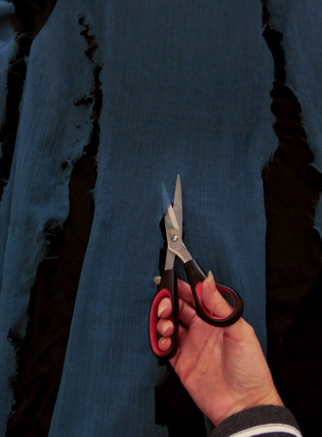 Eine Hand hält eine Schere und schneidet einen Streifen blauen Stoffes mit ausgefransten Kanten auf schwarzem Hintergrund ab – vielleicht, um einen Fetziger Überwurf-Poncho in Übergröße XXXL zu basteln oder einem Hexenkostüm den letzten Schliff zu geben, das sich perfekt als Umhang für Damen und Herren eignet.
