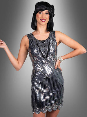 Eine Frau, die das „Premium Paillettenkleider Ruby“ trägt – ein ärmelloses, silbernes Paillettenkleid im Flapper-Stil der 20er Jahre – sowie ein schwarzes Stirnband, posiert vor einem grauen Hintergrund mit Farbverlauf.