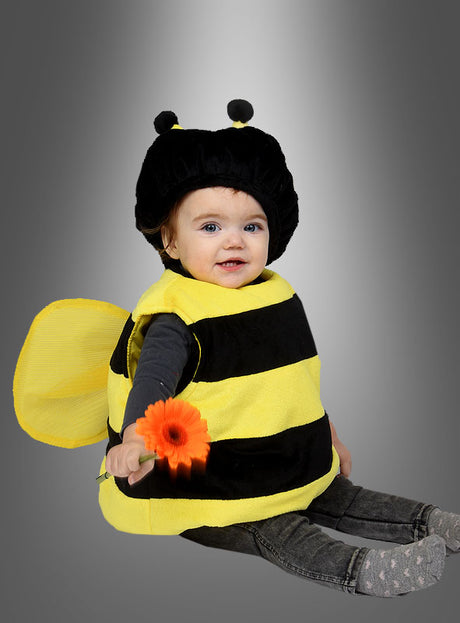 Klar! Hier ist der umformulierte Satz unter Verwendung Ihrer Produktdaten: Ein Baby, das ein gelb-schwarzes, wattiertes Plüschkostüm von „Biene“ trägt, sitzt auf dem Boden und hält eine orangefarbene Blume in der Hand, vor einem grauen Hintergrund mit Farbverlauf.