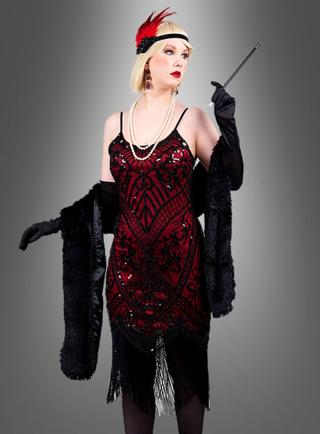 Eine Person, die das „Glamouröses Paillettenkleid Cleo“ trägt – ein rot-schwarzes Flapper-Kleid –, strahlt vor einem schlichten grauen Hintergrund mit schwarzen Handschuhen, einer Perlenkette, einem Haarreif mit Federn und einer Zigarettenspitze klassischen Glamour aus.