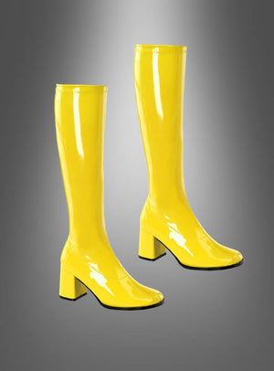 Ein Paar glänzende, gelbe, kniehohe Lackstiefel im Stil der 70er Jahre mit Blockabsätzen, inspiriert von der Mode der 70er Jahre, wird vor einem grau verlaufenden Hintergrund präsentiert. Perfekt, um deinen Disco-Kostüm-Look abzurunden.