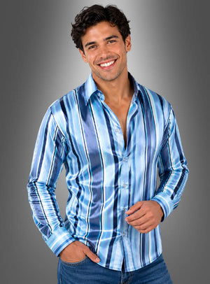 Ein Mann lächelt, während er in einem glänzenden, langärmeligen „Gestreiftes Samt Party Hemd Herren“ mit vertikalen blau-weißen Streifen und einer blauen Jeans vor einem schlichten grauen Hintergrund mit Farbverlauf posiert.