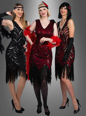 Drei Frauen in Flapper-Kleidern mit Fransen im Stil der 1920er Jahre – eine strahlt im „Exklusives 20er Flapper Kleid Vivian“, eine andere funkelt in einem Paillettenkleid – tragen Stirnbänder, Federboas und hohe Absätze, während sie gemeinsam vor einem grauen Hintergrund posieren.
