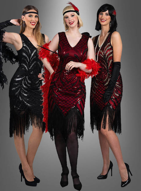 Drei Frauen in Flapper-Kleidern mit Fransen im Stil der 1920er Jahre – eine strahlt im „Exklusives 20er Flapper Kleid Vivian“, eine andere funkelt in einem Paillettenkleid – tragen Stirnbänder, Federboas und hohe Absätze, während sie gemeinsam vor einem grauen Hintergrund posieren.