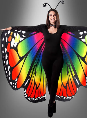 Eine Person, die einen schwarzen Catsuit-Overall und regenbogenfarbene Schmetterlingsflügel trägt, steht mit ausgestreckten Armen vor einem grauen Hintergrund mit Farbverlauf.