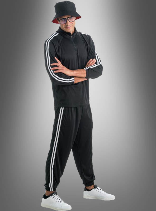 Ein Mann, der einen Rapper-Trainingsanzug mit weißen Streifen trägt, dazu weiße Sneaker, einen schwarzen Bucket-Hat und eine Brille, steht mit verschränkten Armen vor einem grauen Hintergrund mit Farbverlauf.