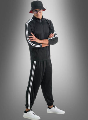 Ein Mann, der einen Rapper-Trainingsanzug mit weißen Streifen trägt, dazu weiße Sneaker, einen schwarzen Bucket-Hat und eine Brille, steht mit verschränkten Armen vor einem grauen Hintergrund mit Farbverlauf.