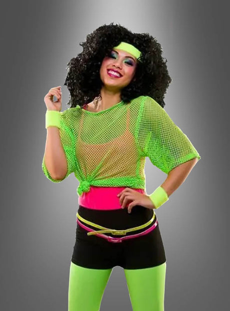 Eine Frau in Sportkleidung im Stil der 1980er Jahre präsentiert ein neonfarbenes, waschbares Netzshirt im Stil der 80er Jahre mit pinkfarbenen Akzenten, schwarzen Shorts und einem Stirnband und lächelt, während sie vor einem grauen Hintergrund mit Farbverlauf posiert – ideal, um einen authentischen 80er-Jahre-Party-Kostüm-Look zu kreieren.