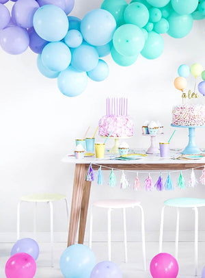 Hochwertige Luftballons in Pastellfarben schmücken einen Partytisch mit Kuchen, Bechern, Tellern und Hockern. Auf dem Boden liegen verstreut Luftballons, während vom Tisch eine Quastengirlande herabhängt.