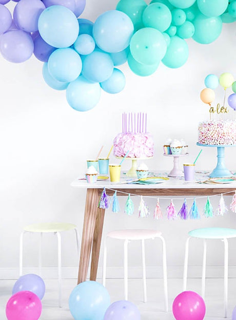 Hochwertige Luftballons in Pastellfarben schmücken einen Partytisch mit Kuchen, Bechern, Tellern und Hockern. Auf dem Boden liegen verstreut Luftballons, während vom Tisch eine Quastengirlande herabhängt.