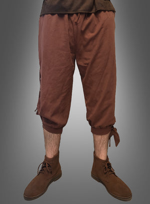 Eine Person, die eine „Mittelalter-Hose für Herren“ trägt – eine braune, kurze Hose, die von LARP-Hosen inspiriert ist und an den Waden mit Bändern gerafft wird –, kombiniert mit braunen Stiefeln, vor einem grauen Hintergrund mit Farbverlauf.