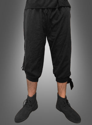 Diese Person trägt die „Mittelalter Hose Herren“ – eine schwarze, knielange Hose, die am Knie gebunden ist – sowie schwarze Stiefel und steht vor einem grauen Hintergrund mit Farbverlauf.