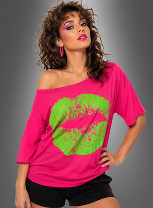 Eine Frau mit lockigem Haar trägt das 80er-Jahre-Shirt „Kiss“ – ein übergroßes Top mit freien Schultern in leuchtendem Pink und einem großen neongrünen Kuss-Aufdruck –, dazu schwarze Shorts, Creolen und auffälliges Make-up; sie steht vor einem grauen Hintergrund mit Farbverlauf.