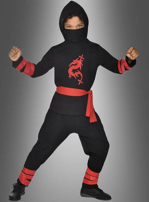 Ein Kind im „Ninja Kinderkostüm rot oder schwarz“ in klassischem Schwarz, mit einem leuchtend roten Drachenemblem, einem passenden roten Gürtel und auffälligen roten Details, nimmt vor einem grauen Hintergrund mit Farbverlauf eine Kampfpose ein – ideal für den Kinderfasching oder jede andere Kostümveranstaltung.