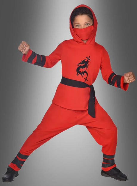 Ein Kind, das das „Ninja-Kinderkostüm rot oder schwarz“ mit schwarzem Drachenaufdruck trägt, posiert mit angewinkelten Armen und Beinen vor einem grauen Hintergrund mit Farbverlauf – damit ist es die ideale Wahl für Kinderfasching-Feiern.