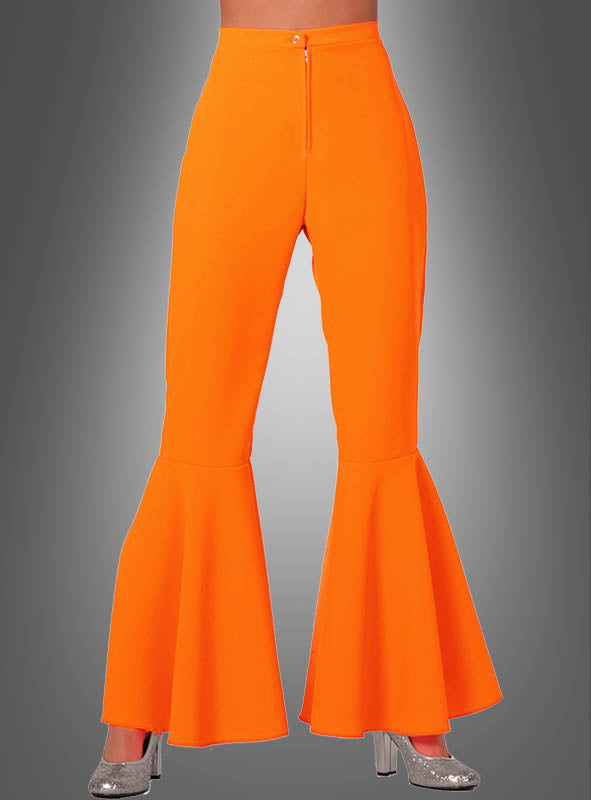 Eine Person, die die Neon-Schlaghose „Damen Schlagerstar“ in leuchtendem Orange und silberne Glitzer-Plateauschuhe trägt, steht vor einem grauen Hintergrund mit Farbverlauf.