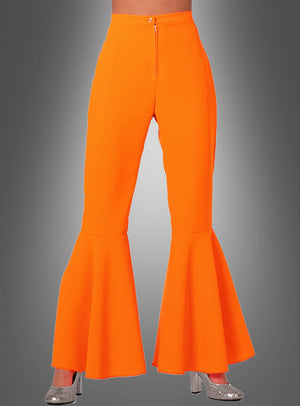 Eine Person, die die Neon-Schlaghose „Damen Schlagerstar“ in leuchtendem Orange und silberne Glitzer-Plateauschuhe trägt, steht vor einem grauen Hintergrund mit Farbverlauf.